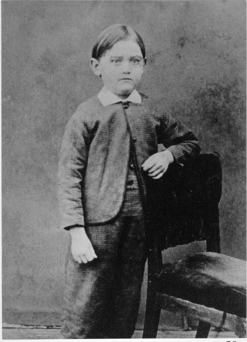 En liten pojk, A E Bolling, i helfigur. Han är klädd i smårutig jacka, väst och byxor. Han vilar vänster arm på en stolsrygg.