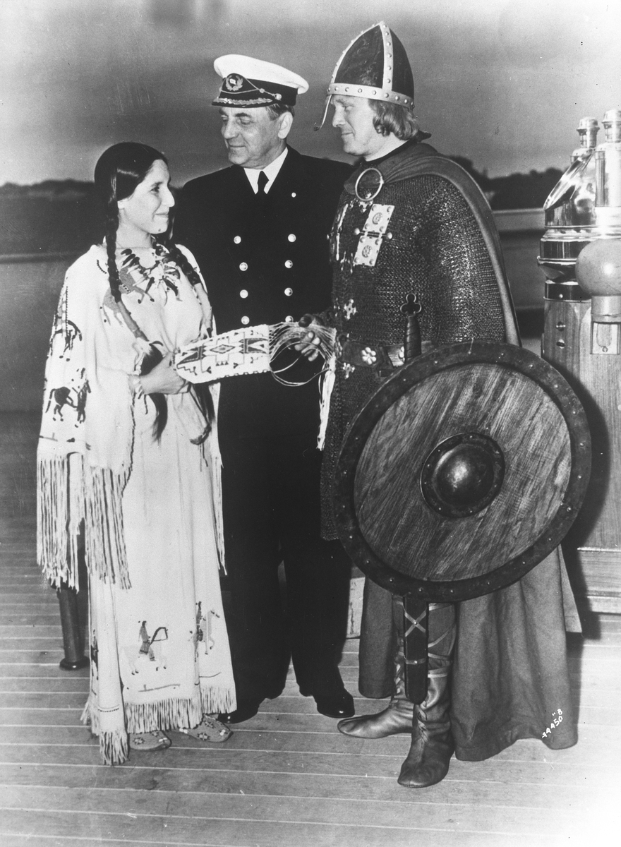 Ombord på Den Norske amerikalinje. En viking og en indianerkvinne overrekker hverandre gaver, kaptein Ingens står mellom.