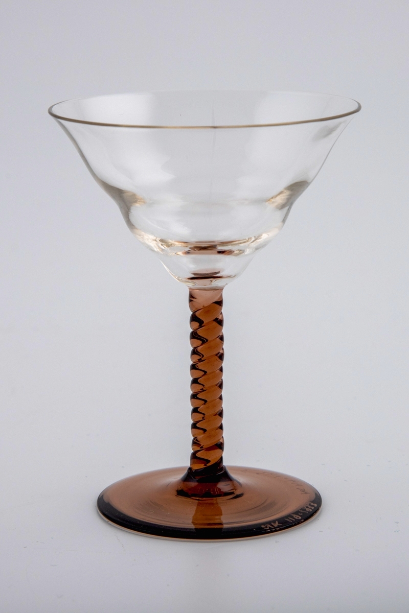 Cocktailglass med kupa i klart glass. Fot og og stett er utført i gjennomskinnelig brunfarget glass. Sistnevnte består av to vridde glasstaver i spiralform.