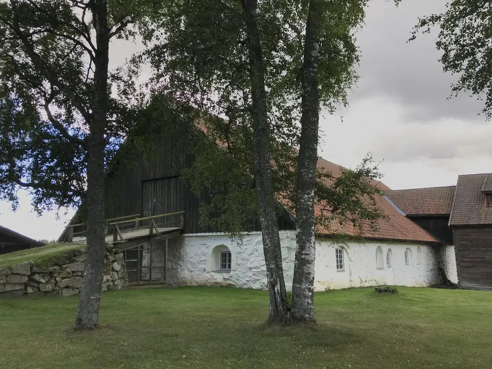 Bildet viser et fjøs. Foran fjøset er det to store trær som delvis skjuler fjøset. Fjøset er brunt og hvitt.