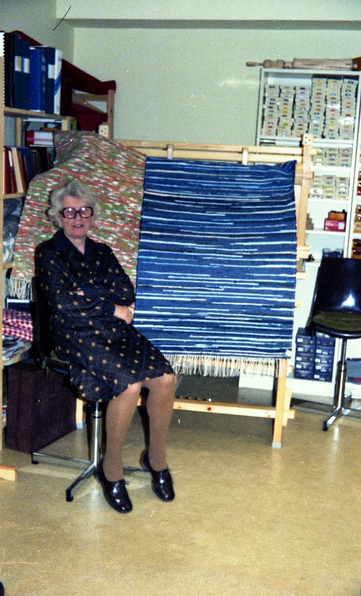 Julförsäljning av hantverk i pensionärernas hobbylokal på Våmmedalsvägen år 1977. En kvinnar sitter brevid en vävstol där det hänger två handvävda mattor.