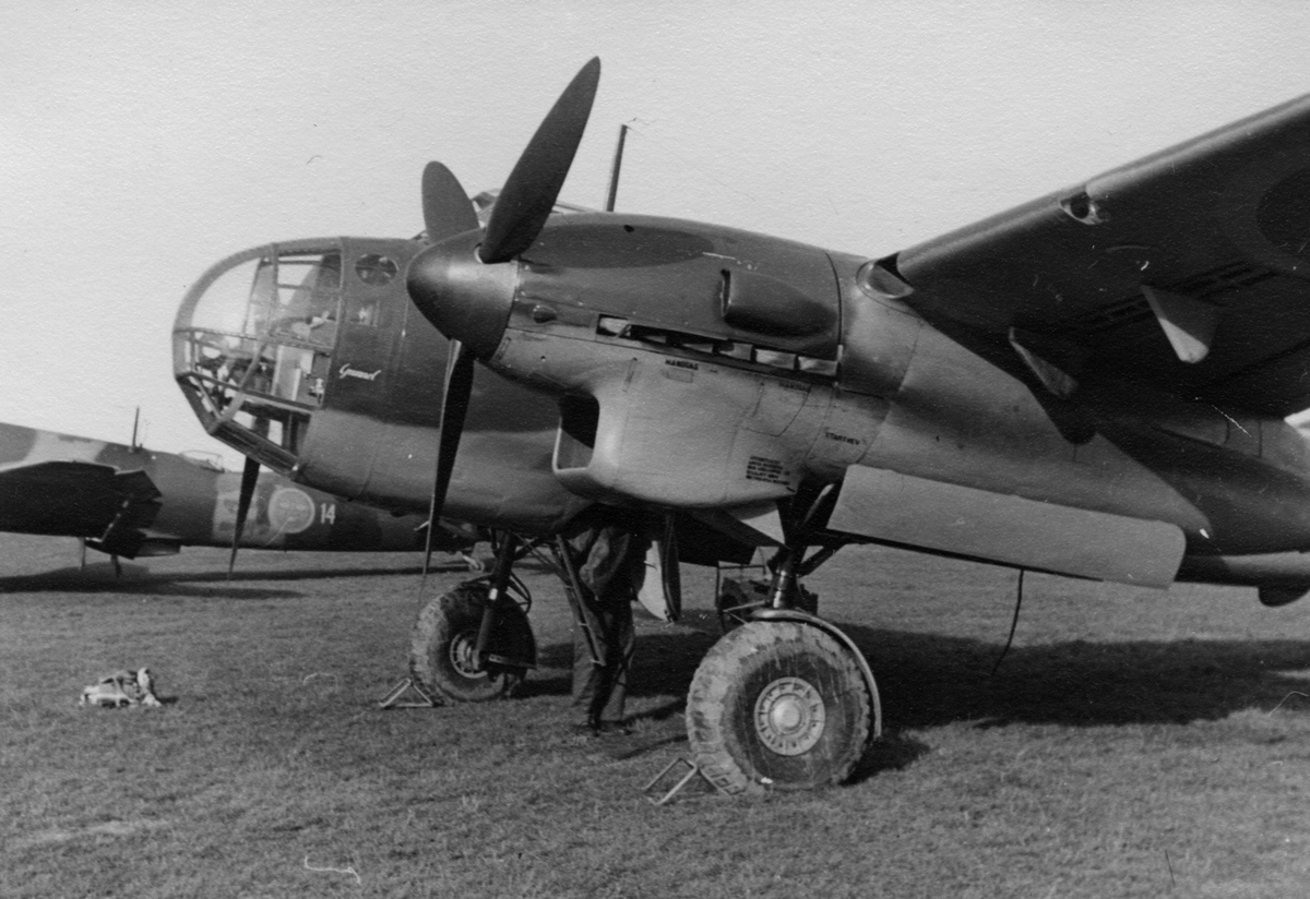 Flygplan Saab B 18 står på ett flygfält, 1940-tal. En person arbetar med flygplanet. Flygplanet är märkt 'Gunnel'. I bakgrunden står ett flygplan B 3 Junkers Ju 86K tillhörande F 14 Hallands flygflottilj.