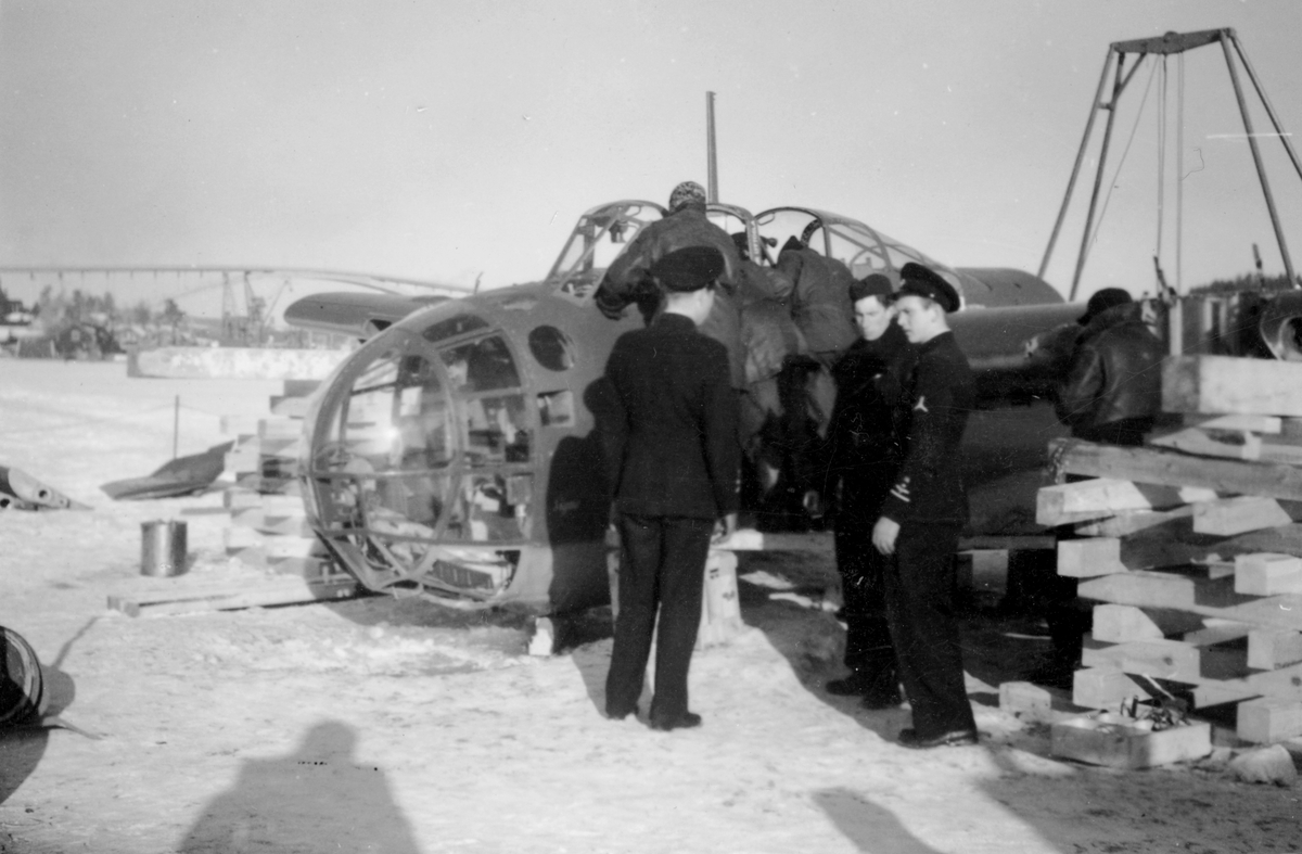 Flygtekniker och flygvapenpersonal samlade vid bärgat flygplan B 18 på kajen i Härnösands hamn efter nödlandning på isen den 10 februari 1946. Motorerna har demonterats. I bakgrunden syns en lyftkran som användes vid bärgningen.
