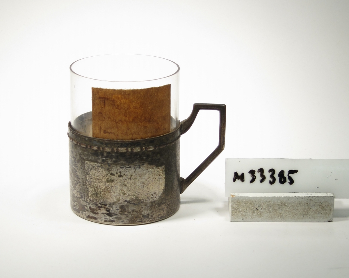Tekopp, cylinderformad, med tillhörande hållare i metall.
Lapp: "Theglas
Bry???
1 cm lägre (???)"