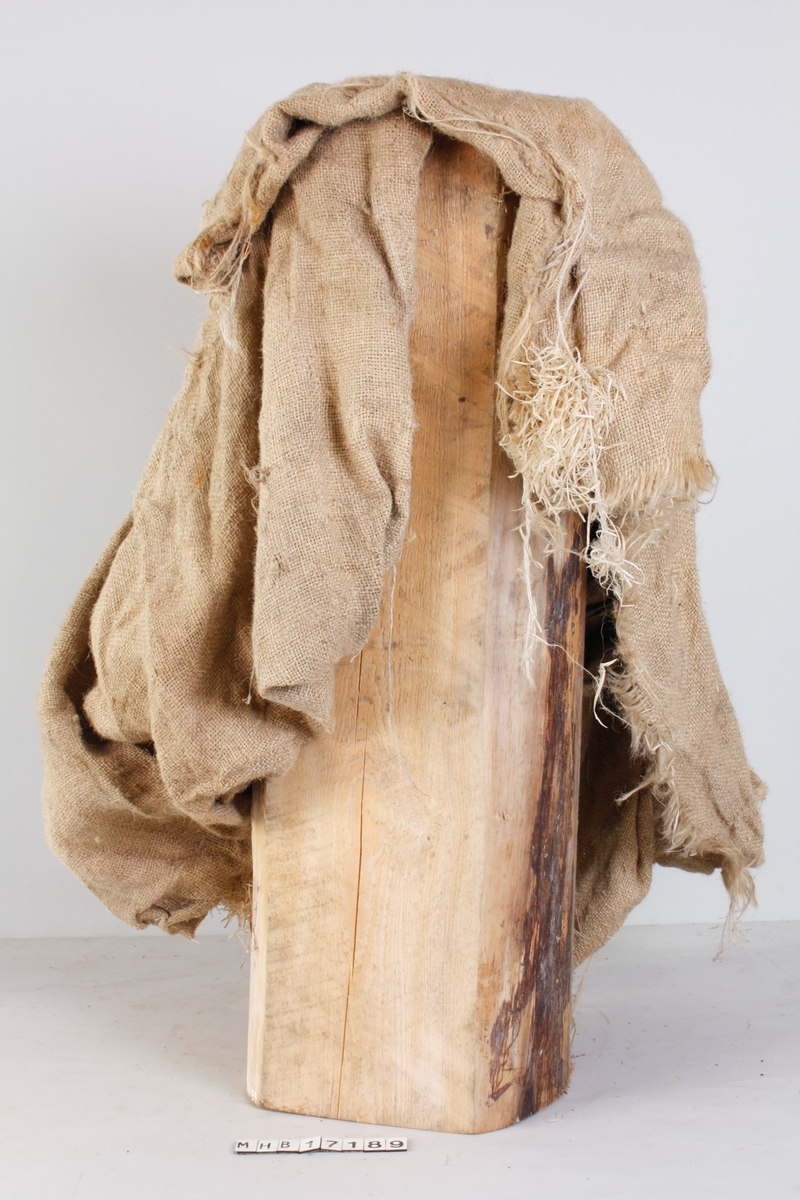 Trestubbe/kubbe, en del av en barket tømmerstokk. Over stokken ligger en grovt vevet tekstil. Ukjent bruk.