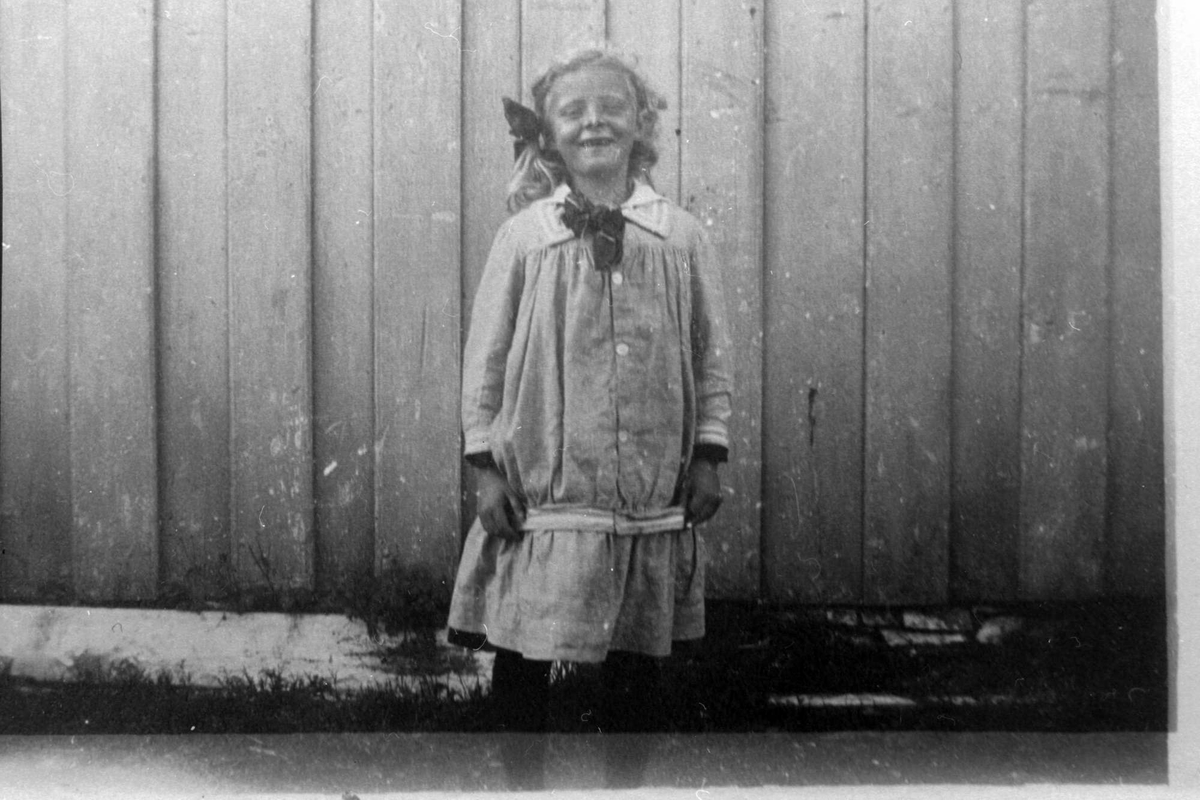 Amatørportrett av jente med knekort kjole og sløyfe i håret stående ved husvegg.