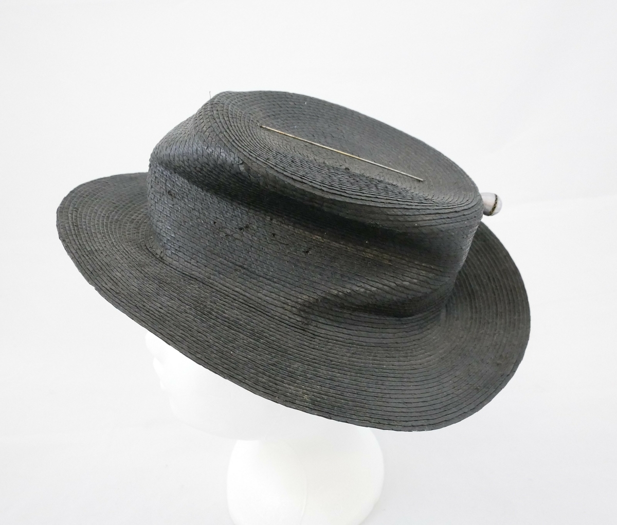 Sort stråhatt med flat pull og rund brem. Den er foret med sort bomullsstoff og på toppen av hatten er det en hattenål. 