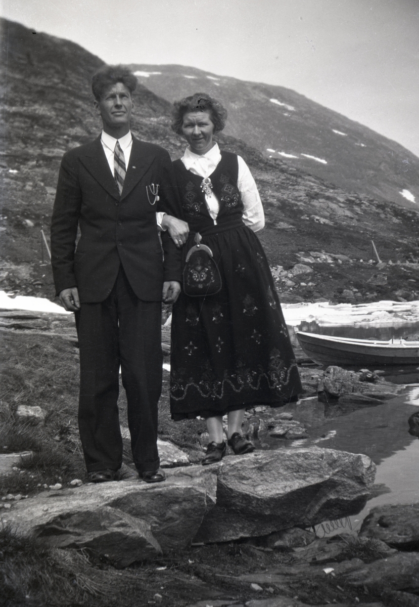 Portrettfotografi av en mann og ei kvinne med dress og bunad. I bakgrunnen  ligger en robåt på et vann.
