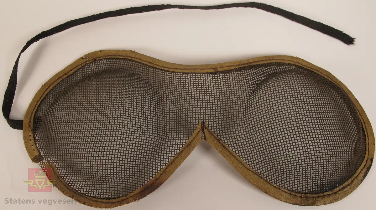 Vernebriller av netting i metall, med påsydd tekstil rundt kanten. Det er sydd på strikk på hver side av vernebrillen for festing rundt hodet, men strikken har falt av på den ene siden.