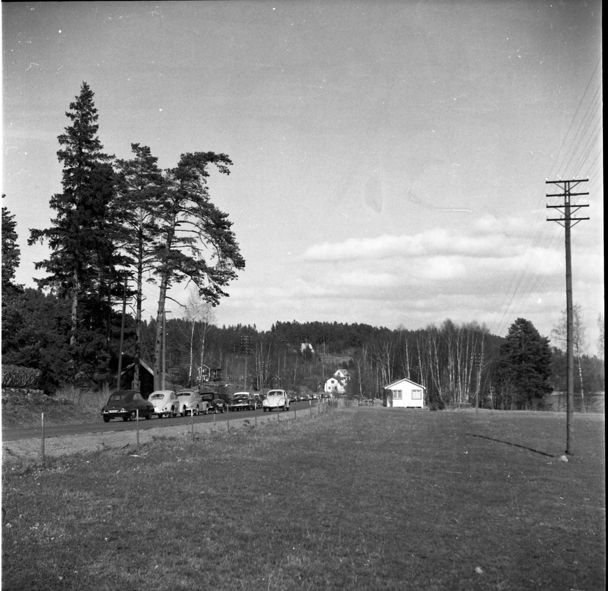 En lång rad bilar står parkerade på Tranåsvägen vid Bunnström med badplatsen till höger bakom björkdungen. En VW kommer körande mot fotografen. Enstaka hus utmed vägen.