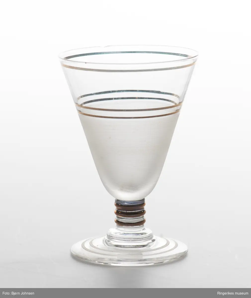 Fire akevittglass dekorert med tre gullstriper øverst på glasset tre tykkere gullstriper på stetten. Nederste del av glasset er etset og foten har to tynne etsete striper.