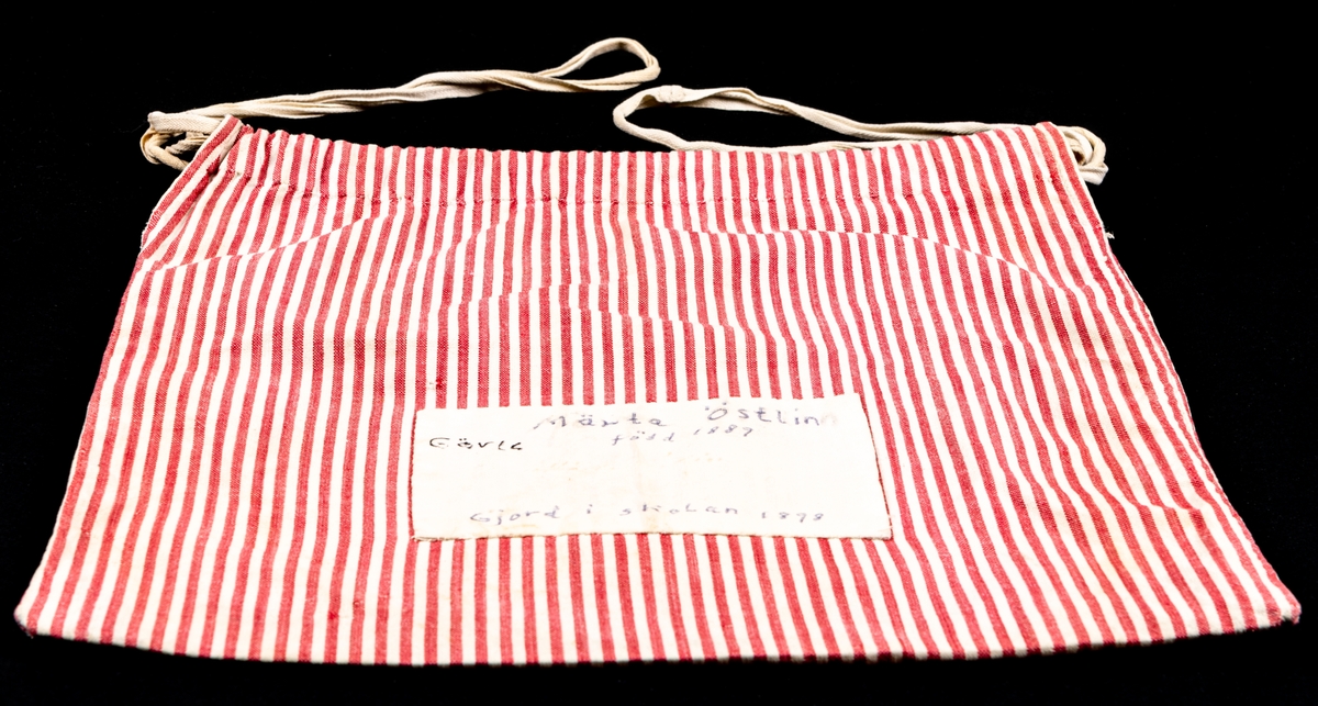 Handsydd tygpåse av röd, vitrandigt bomullstyg, maskinvävt tuskaftat. Upptill en dragsko med ett vit bomullsband. På ena sidan finns en vit tyglapp med texten: Märta Östlin Gävle, född 1889 gjord i skolan 1898.