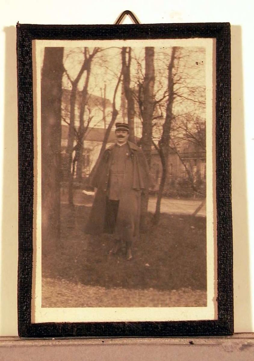 Fotografi; utendørsopptak: en voksen mann i militæruniform med stor lang kappe over skuldrene. I bakgrunnen skimtes en stor bygning og en del trær. 