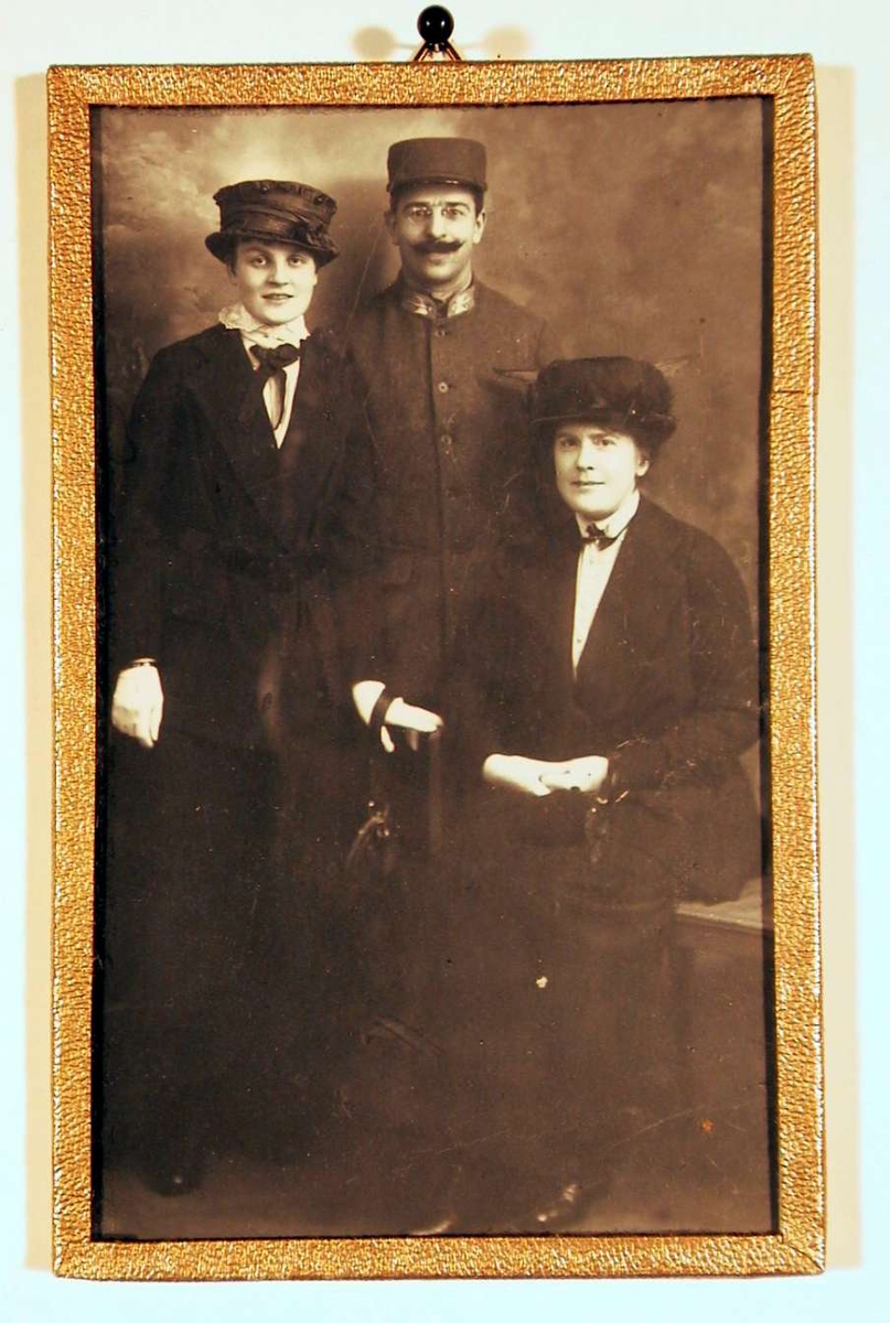 Fotografi av en mann i uniform og to damer, den yngste stående og den eldre sittende på benk i atelier.
Alle ser direkte på betrakteren.