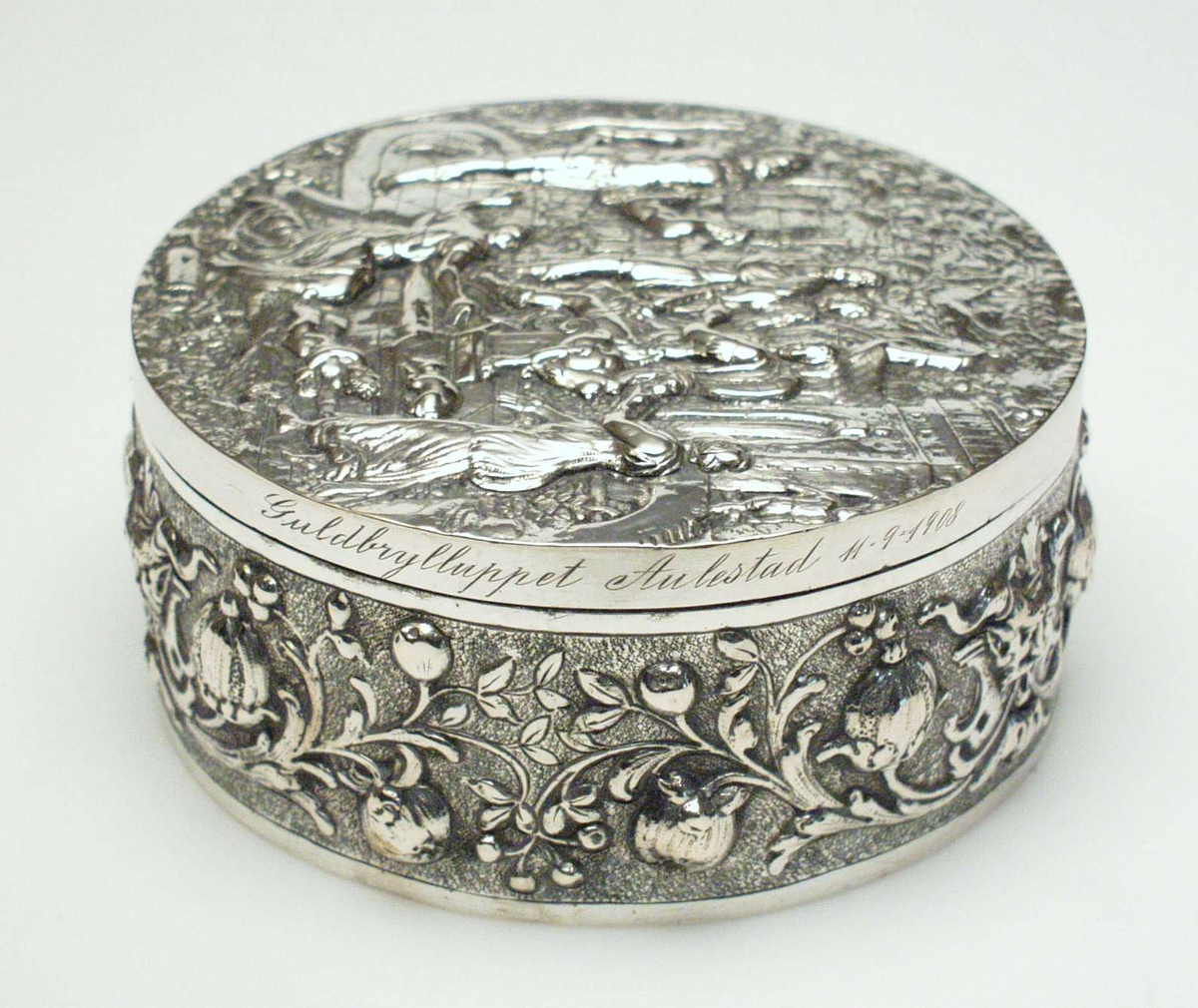 Rundt skrin i sølv med støpt dekor i relieff. På lokket et bibelsk motiv i relieff, på esken et rosemønster