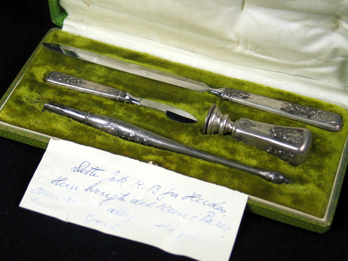 Skrivesett med inngravert monogram. Settet består av en brevkniv, kniv (?), penneskaft og stempel dekorert med blomstermotiv. Det ligger i en grønn eske i kartong fôret med fløyel og silke.
