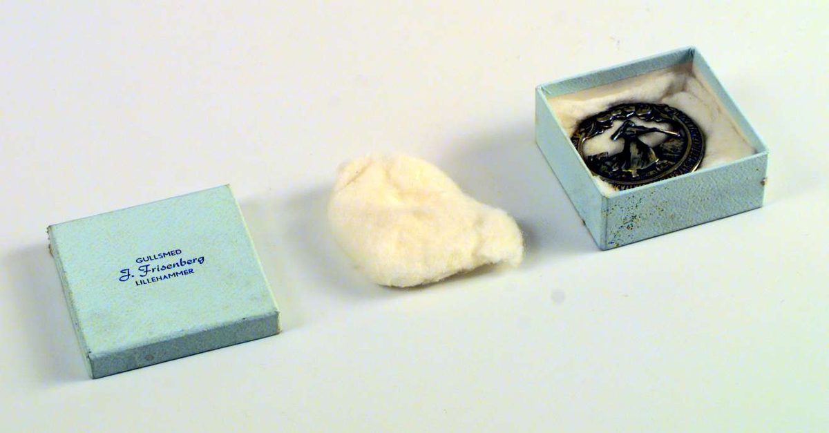 Brystnål eller brosje i sølv med Pillarguri-motiv. Nålen ligger i en eske fra gullsmed J. Frisenberg. Esken er lys blå.