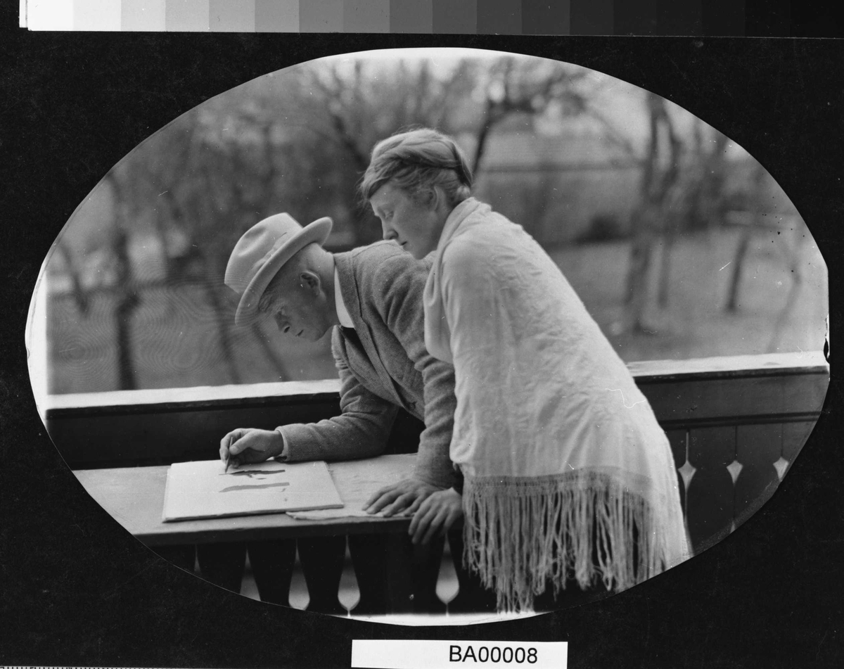 Utendørsfotografi; veranda med mann og dame som lener seg inn over bord, hvor han er i ferd med å tegne.