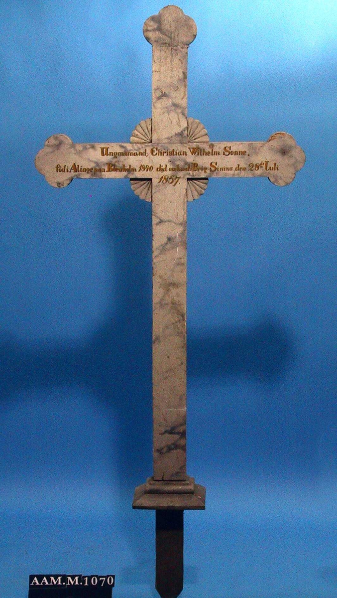 Kors i tre, malt lyst grått med marmorering og innskrift. 