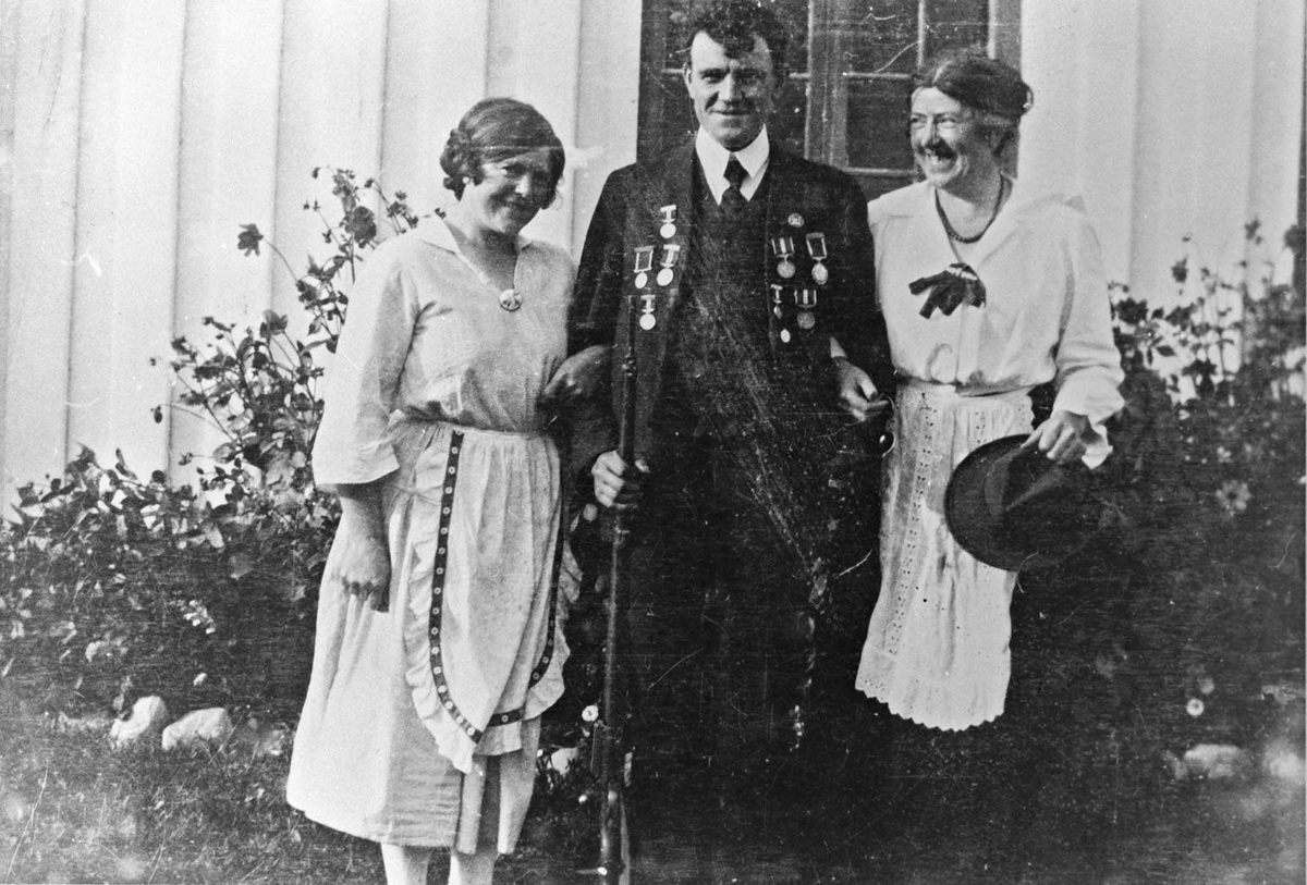 Gruppebilde, 2 kvinner flankerer en mann fotografert i hagen utenfor et hus. Mannen har gevær og jakkeslagene full av medaljer.
