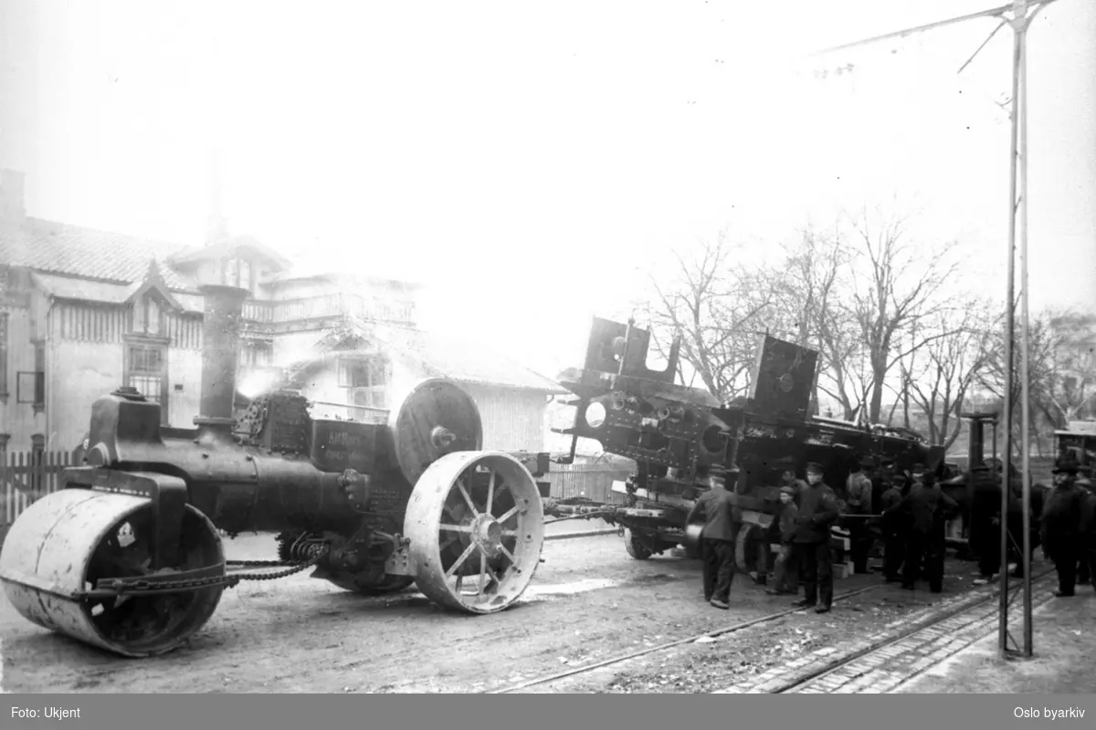 Dampveivals som trekker i damplokomotiv-chassis i gata. Lasten har forskjøvet seg, og det arbeides med å jekke den på plass igjen. Skarpsno (Drammensveien 84) i bakgrunnen. Se også A-20189/U/001/155. Tilbakemelding fra bruker: "Bildet er overveiende sannsynlig tatt før åpningen av Havnebanen i 1907, hvor gatetransporten av lokomotiver opphørte. Eneste lokomotiv av denne typen som Thune leverte før den tiden, var Gjøvikbanens nr. 144. Det ble levert 12. mai 1906, og bildet er derfor sannsynligvis tatt i månedskiftet april/mai 1906."