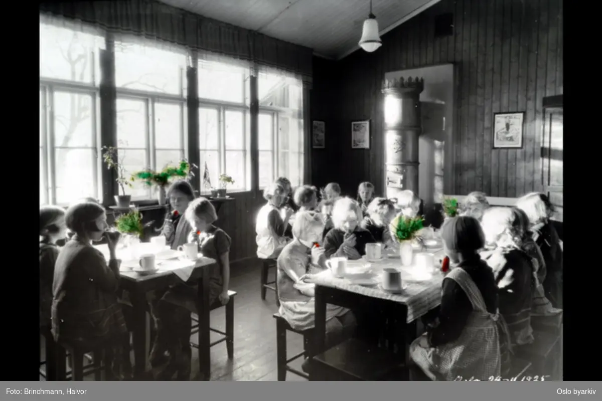 Skoleelever som spiser. Skolefrokost, oslofrokost? Fra Sandaker friluftsskole? Bildet datert 24.11.1925. Håndkolorert (blomster i vinduskarm og på bordene, gulrøtter som jentene spiser)