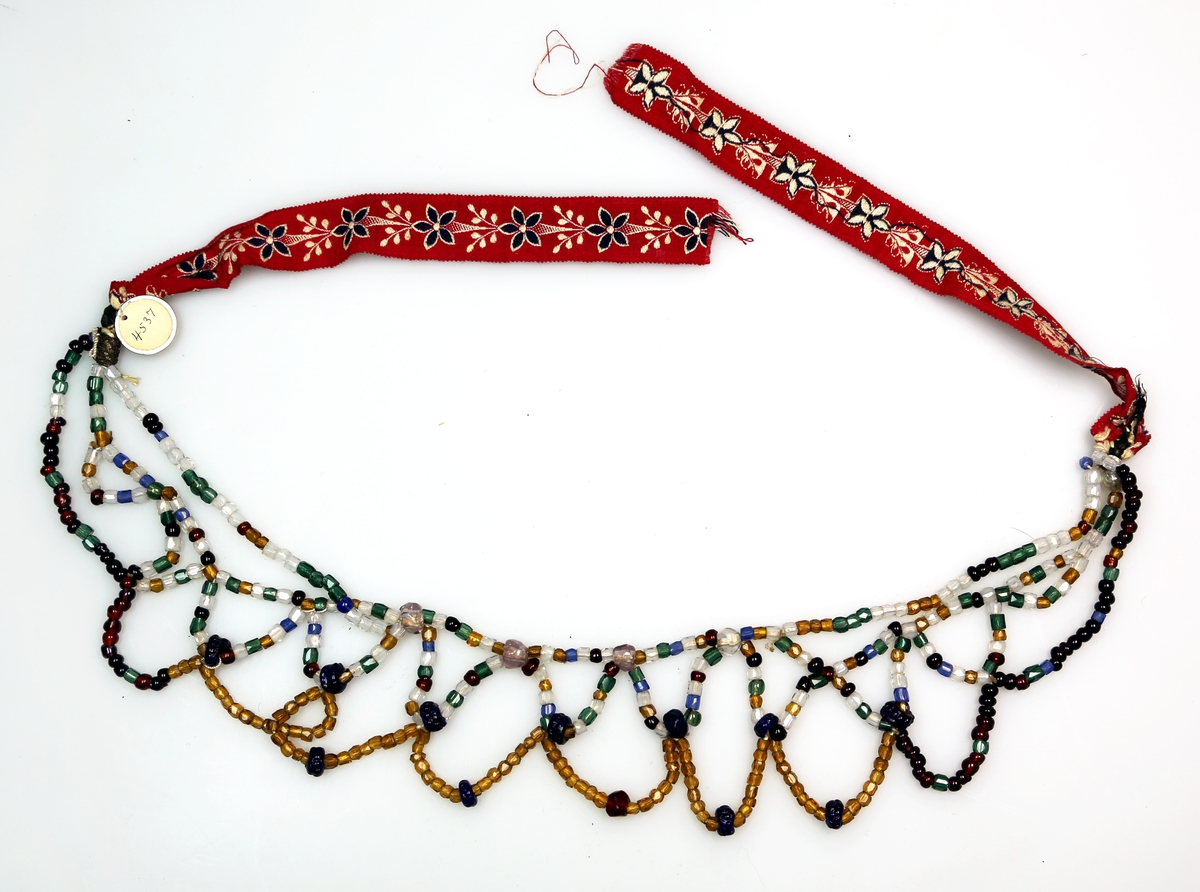 Perler i ulike farger og utforming tredd på snorer, som er knytt i mønster og som har bomullsbånd med blomstermotiv i hver ende til knyting.
