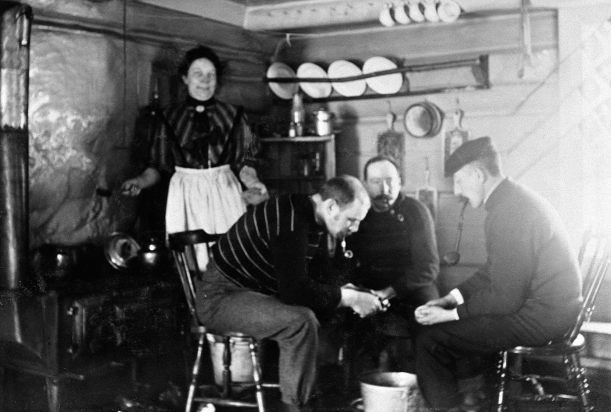 Interiør, kjøkken med vedkomfyr. Tre menn med piper skreller poteter. Dame (kokka) ser på. I midten Philip N. Petersen, Furnesåsen.