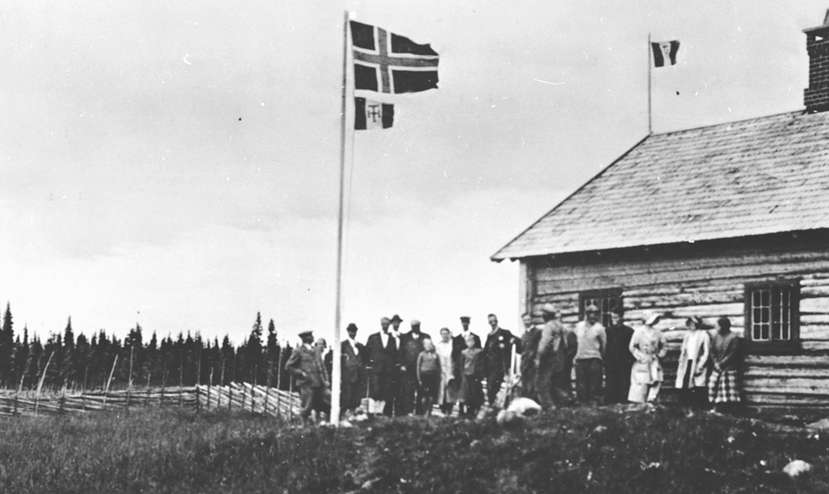 Gruppe samlet utenfor hytte på Gammelskolla seter, Ringsakerfjellet. Hamar og Hedemarken turistforening kvarter. Flagg og vimpel.