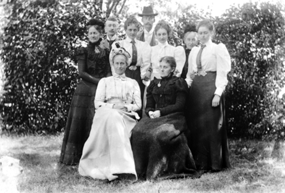 Ni voksne fotografert i hagen på Grefsheim. Sittende fra venstre er Julie Munch Gram og Laura Larsen Naur (F.Munch). Stående er Agnes Petersson (F. Irgens), Mads Gram, Thekla Munch, hoffdame hos dronning Maud, Janken Gram, Lol (Julie) Nissen, f. Mellbye, (1876-1960), Sofie Mellbye (F.Munch), Sofie Pihl (F.Mellbye).