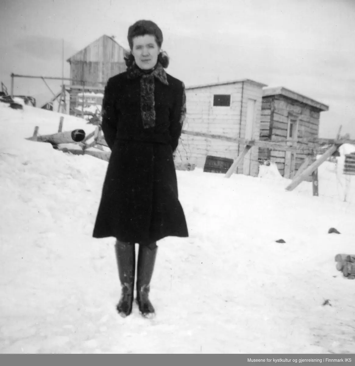 Rikarda Kristensen i kåpe og støvler. Arthur Ingebrigtsens hus i bakgrunnen.
Midt på bildet et utedo, ca 1954.