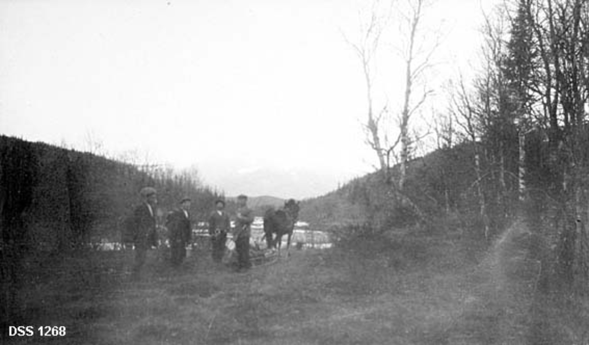 Fotografiet viser fire karer med ryggsekker og spann, samt en hest med slede på barmark ved ei elv.  Omkring dem står bladlaus bjørkeskog, i bakgrunnen skimtes et fjell med snøflekker. 