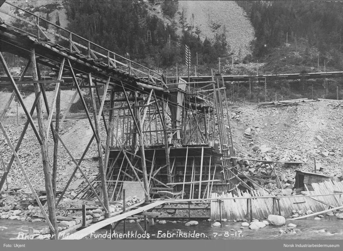 Bygging av fundamentkloss for Birkelandsbroa på sørsiden av Måna. Broen har fått en midlertidig transportbro fra nord- til sørsiden. Trakt fra transportbroen for å få masse ned til fundamentet. Skur bygget av bølgeblikk satt opp på elvebredden vest for broen. Rørledninger fra ovnshuset på Såheim i fjellsiden.