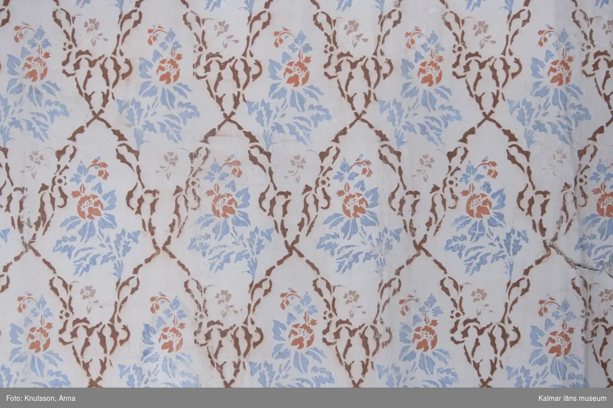 KLM 39750. Tapet, schablontapet. Schablonmålad tapet med vit bakgrund, röda blommor, blått bladmönster och brunt snedrutemönster. Ljusblå bård med röda blommor och vita blad bredvid varandra. Datering, 1840-tal.