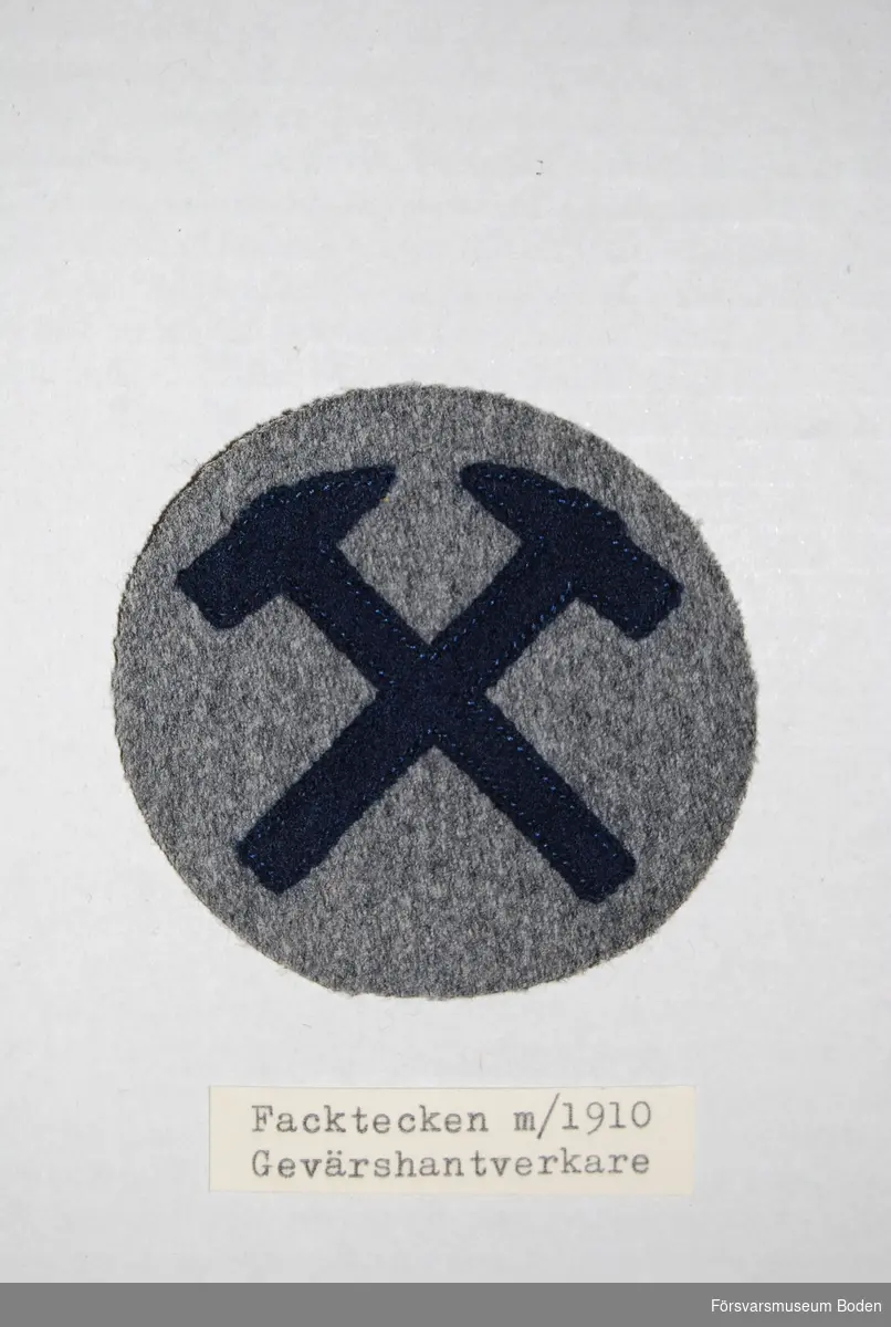 Två korslagda hammare av mörkblått kläde på grå bakgrund. Syddes fast på uniformens högra underarm.