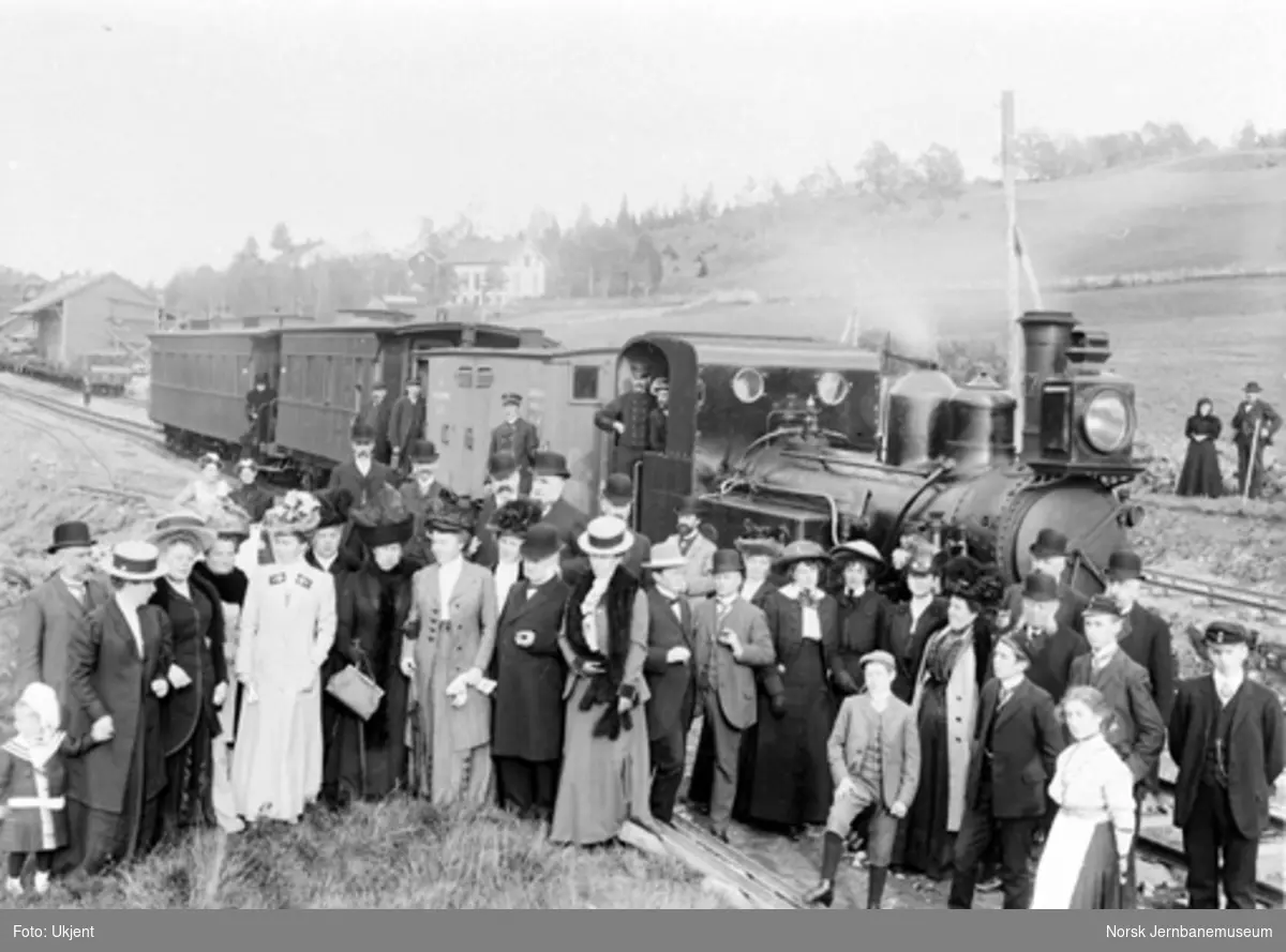 Jernbanepersonale og innbudte gjester oppstilt for fotografering foran toget på Åmli stasjon, sannsynligvis i 1910. Damplokomotiv type IV nr. 17 "Vegtam" foran toget