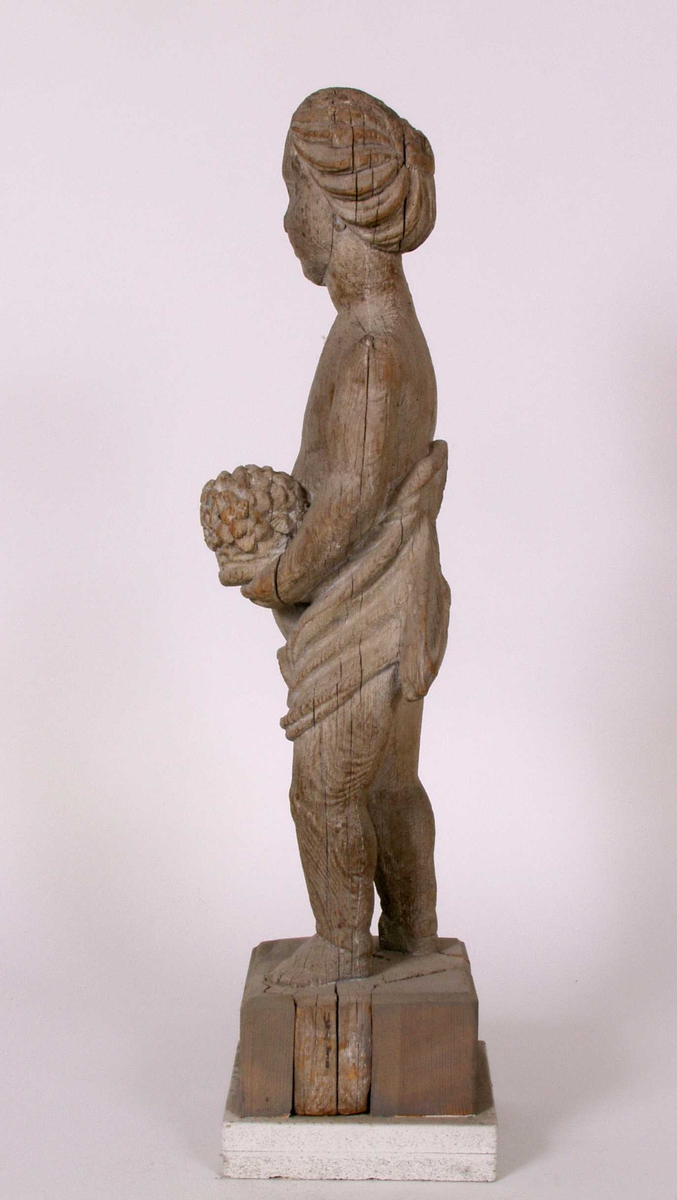 Utskåret trefigur. Det finnes rester av hvit maling i fordypninger (anatkelig linoljemaling. Dertil finnes det et tynt gråhvitt malingslag over hele overflaten. Dette lag er sekundært ettersom det også er påført der deler mangler.