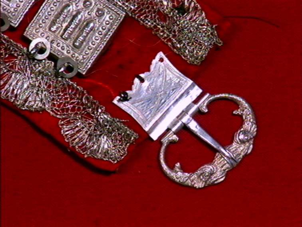 Rødt belte med sprette og sprot.
Stølar av sølv på rødt klede, sølvbeslag og metallknipling.