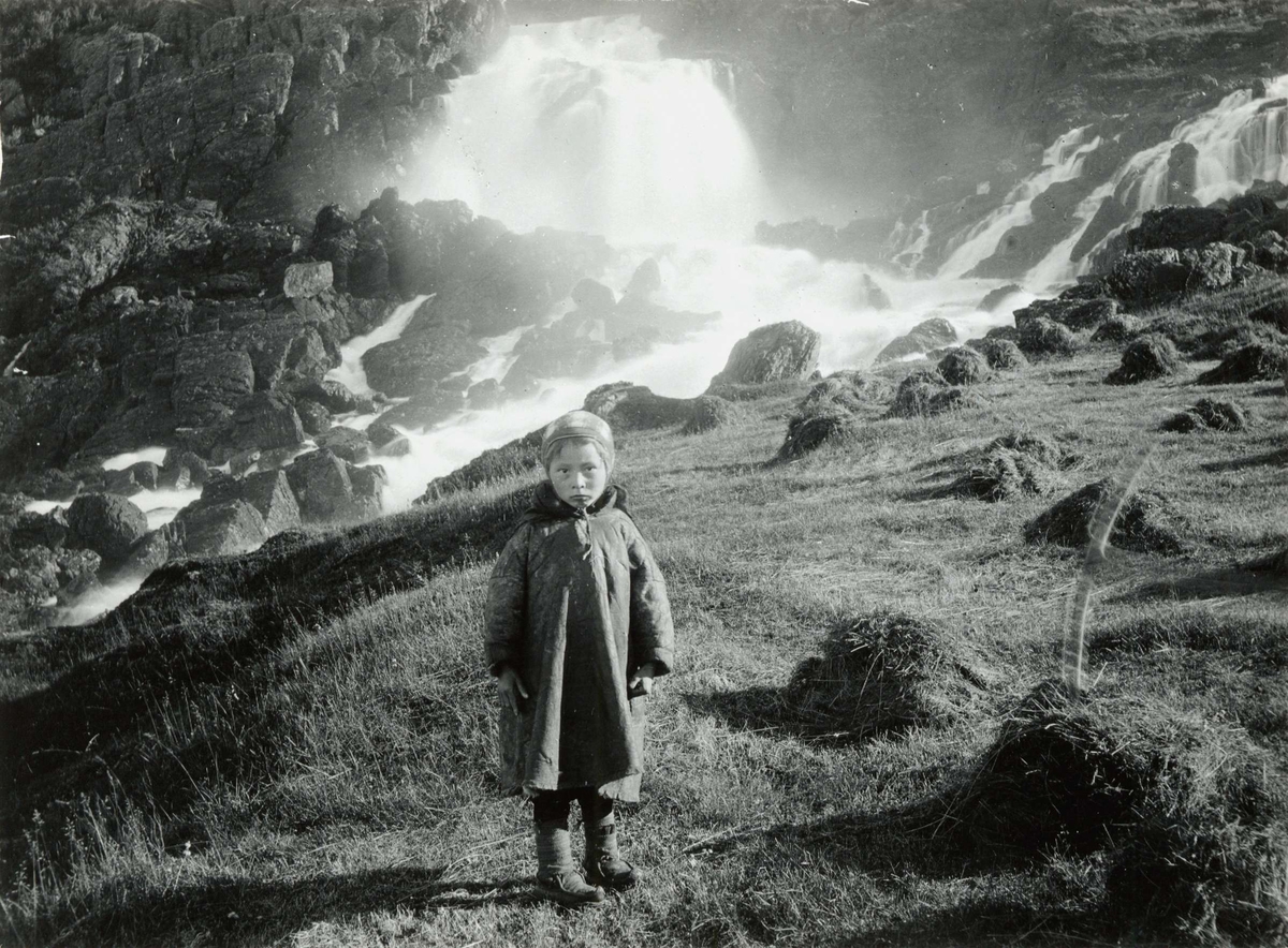 Pike i samisk saueskinnskofte, Valborg 8 år, stående foran Adamsfjordfossen, Lebesby. 
Del av serie fra en forskningsreise i Øst-Finnmark 1909.