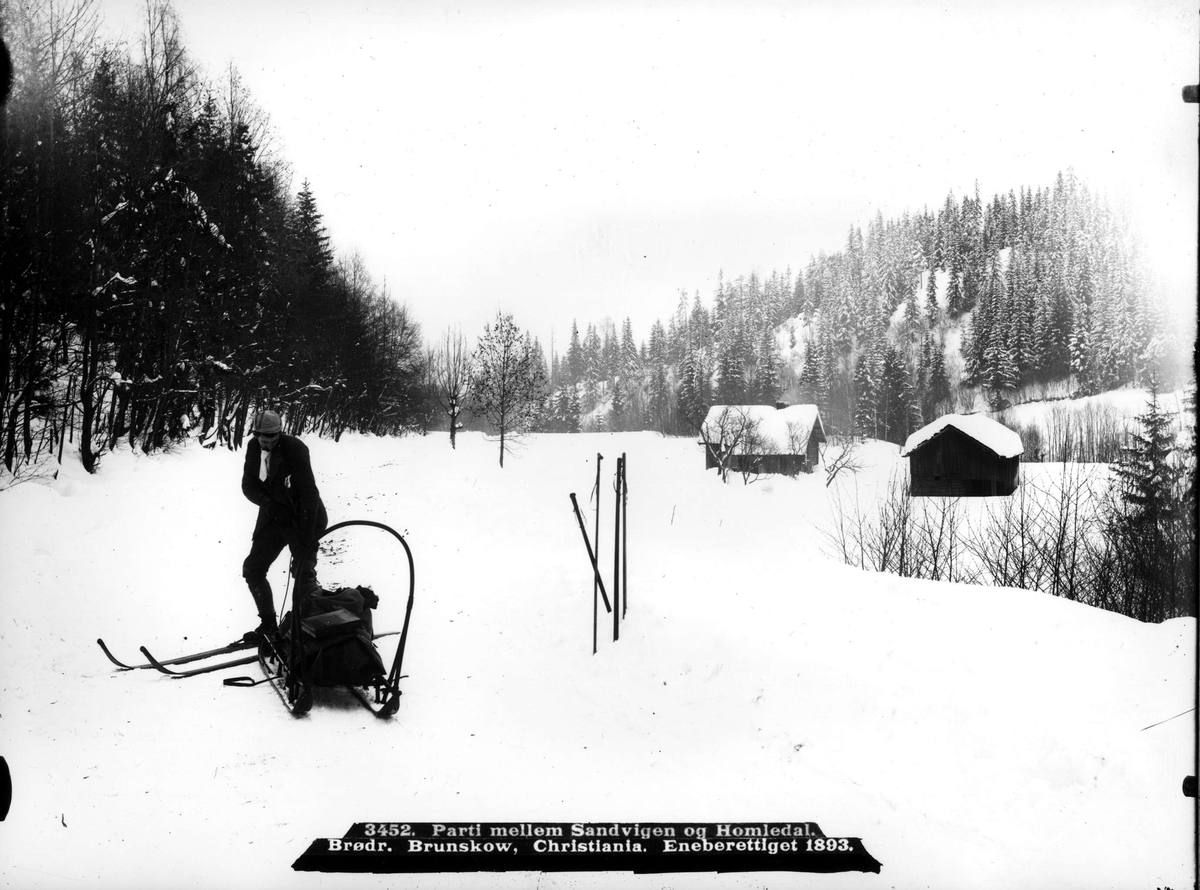 Skiløper med slede med oppakning  i snørikt landskap med to hus. Ant. en av fotografene Brødrene Brunskow. Del av deres serie norske landskapssbilder.
