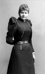 Portrett, kvinne i svart kjole med belte. Brosje i halsen. F