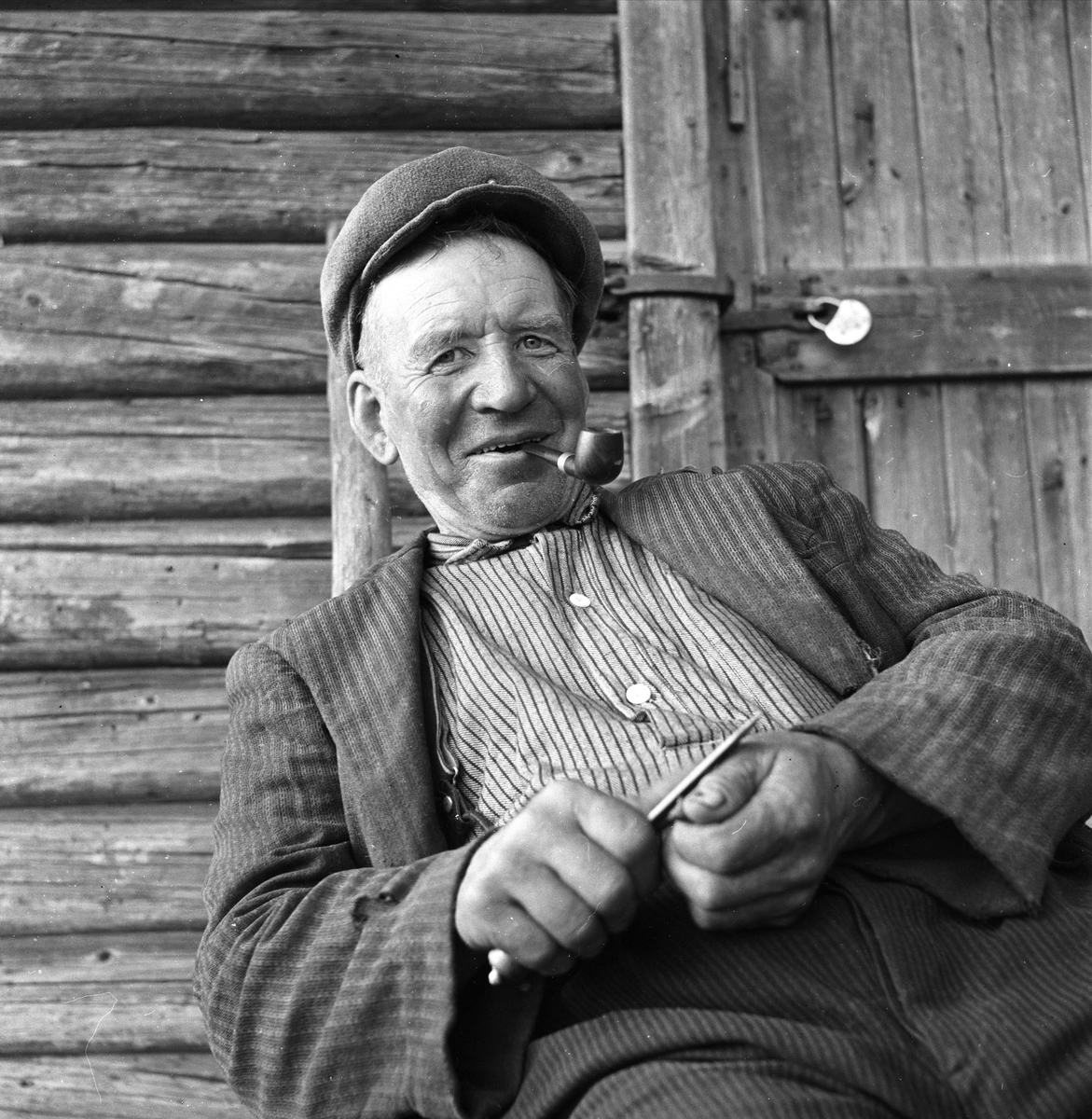 Tømmerfløtere i Sperillen, Ringerike, Buskerud, ant. 1950-tallet. Portrett av mann med pipe i busserull som sitter og spikker.