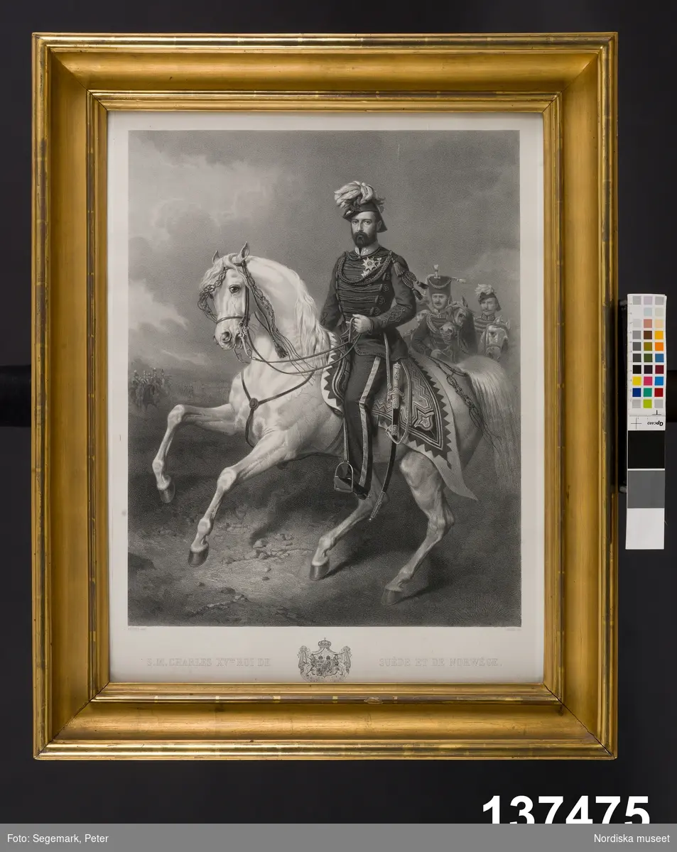 Katalogkort:
"Porträtt i litografi, framställande Karl XV till häst, i förgylld ram med kunglig krona. 1800-talets andra hälft. Tryckt text: 'S.M. CHARLES XVÈME ROI DE SUÈDE ET DE NORWÈGE' 'KIÖRBOE pinxt LAFOSSE lith'. Handskriven lapp på baksidan med historik över kungen samt : 'Taflan inköptes på auktion den 26 mars 1907 i Helsingborg efter aflidne kammarherren hos Drottning Lovisa, Henrik af Hammareus /?/ som Carl XV ofta benömnde Henrik Fogelfängaren under glada stunder. O.K". kronan på ramen saknas 1990. AT/SEN 1990" [Annika Tyrfelt, Sigrid Eklund]