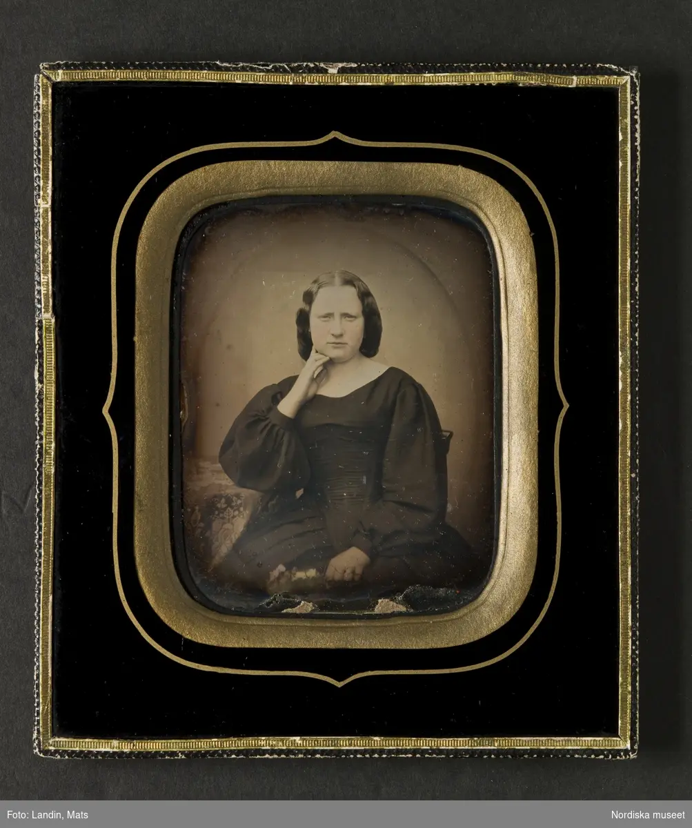 Handkolorerat porträtt av sittande ung kvinna iklädd mörk klänning med vid kjol, puffärmar, mittbenat, uppsatt hår. Höger hand under hakan, vänster hand håller en liten blombukett. Dagerrotyp / daguerreotyp, 1850-tal. Nordiska museet inv.nr 249999.
-
Portrait of an unidentified young woman. Sixth-plate tinted daguerreotype, 1850s.