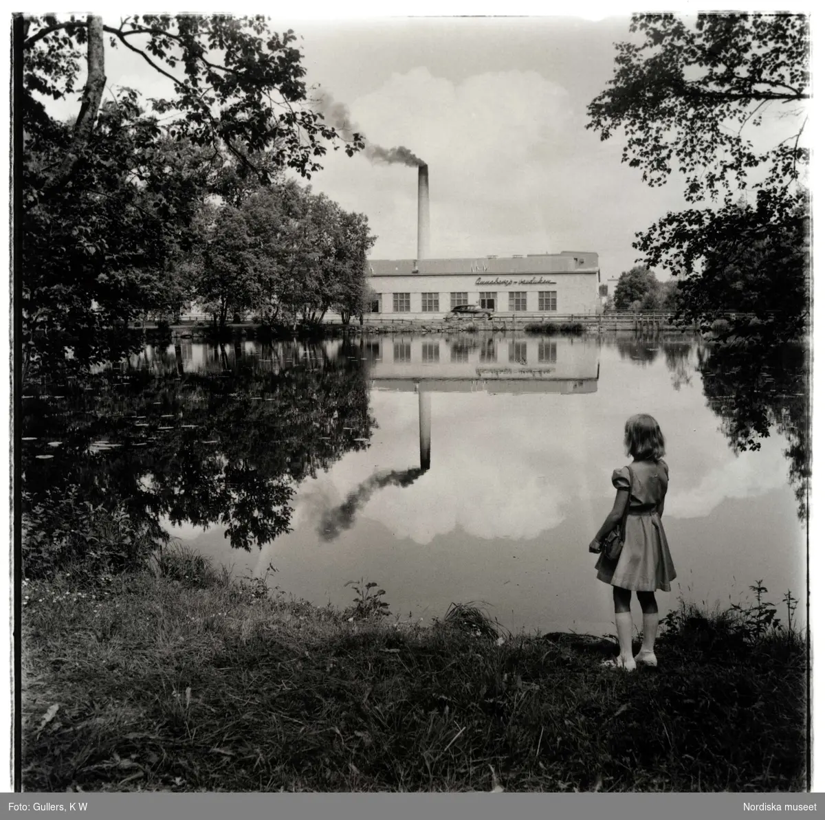 Annebergs Vaxduksfabrik. Exteriör. En flicka står och tittar över vattnet mot fabriken.