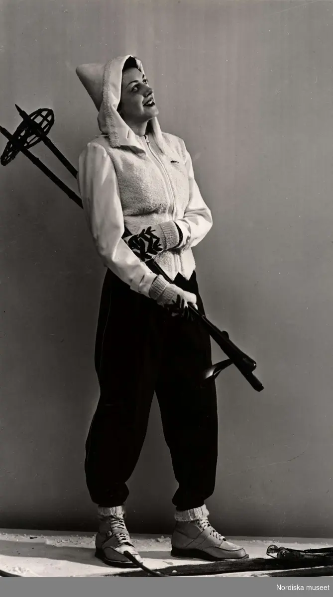 Vintermode. En kvinnlig modell poserar iförd skiddräkt. Hon står brevid ett par skidor och håller ett par stavar i handen. Nordiska Kompaniet 1939.