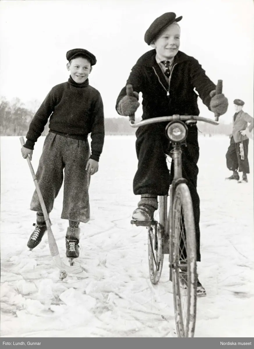 Två pojkar på isen. Den ene åker skridskor och bär en bandyklubba, den andre cyklar.