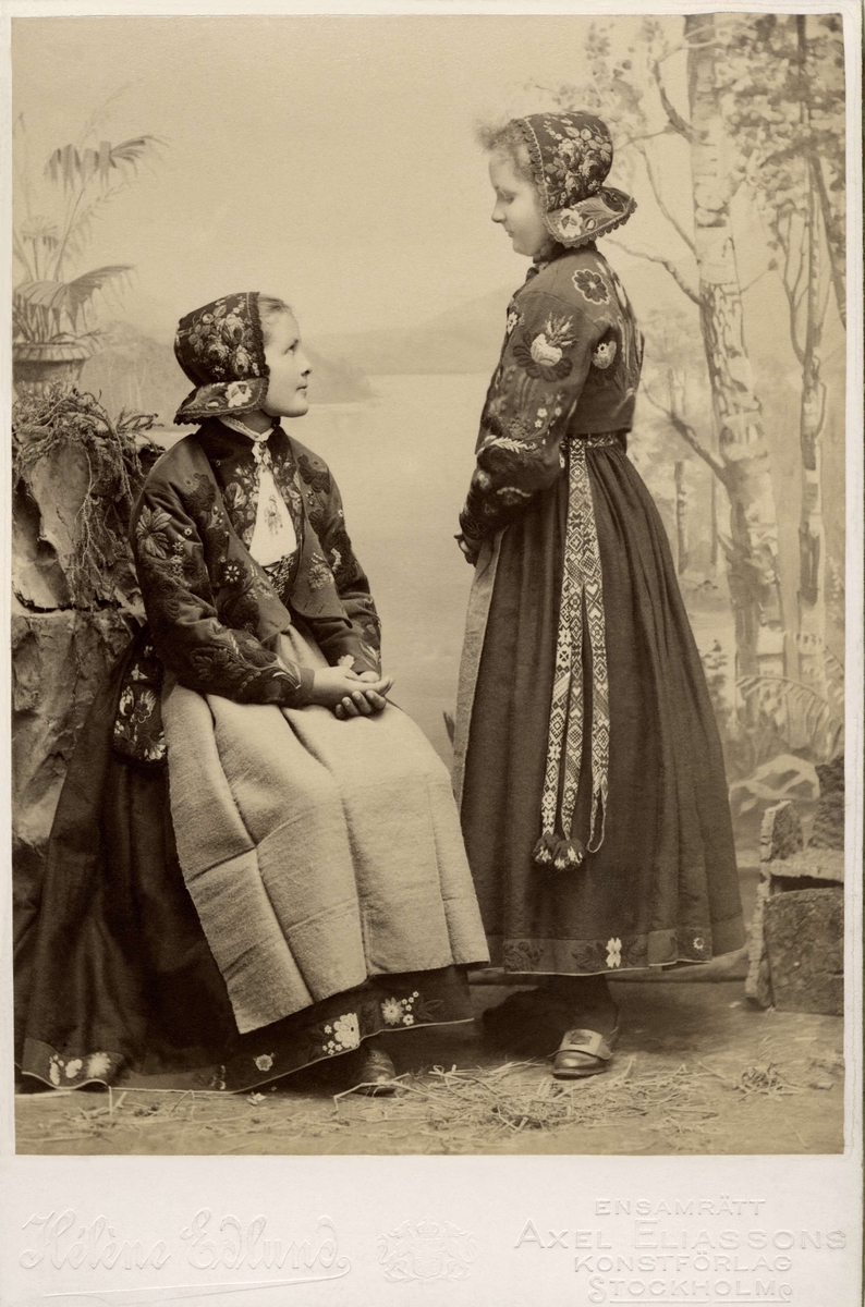Två unga flickor poserar i dräkter från Dala-Floda med blommiga jackor och hättor.