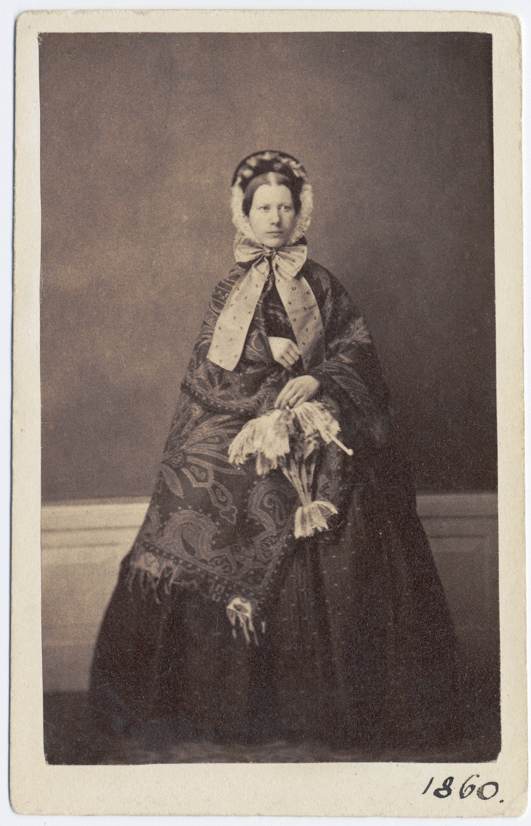 Kabinettkort. Porträtt, helfigur, av fru Thérèse Weber f. Widelius. Hon är iklädd mörk, vid kjol och över axlarna bär hon en stor, paisleymönstrad sjal. Hon har mössa med breda band knutna i rosett under hakan och ett parasoll i handen.