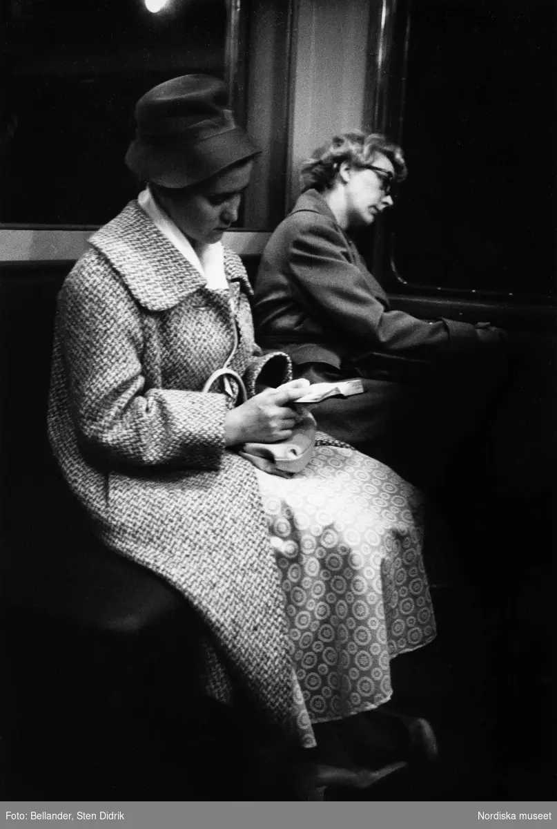 Två kvinnor sitter på tunnelbanan. Den främre är klädd i hatt och kappa och ser ut att titta i sin kalender. Den andra lutar sig mot fönstret som vetter ut mot mörkret.
Stockholm.